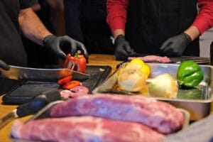 Man erkennt Fleisch, Zwiebeln und Paprika. Eine rote Paprika wird mit einem Messer auf einem schwarzen Brett geschnitten