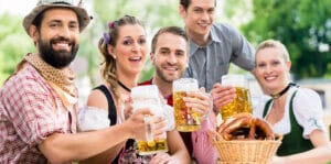 Fünf Personen in traditioneller bayrischer Tracht stoßen mit Bier gefüllten Maßkrügen an; auf dem Tisch steht ein Korb mit Brezeln