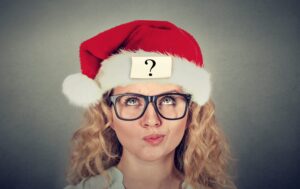 Eine blonde Frau mit Brille schaut fragend nach oben; sie trägt eine Weihnachtsmütze, auf der ein Zettel mit einem Fragezeichen klebt